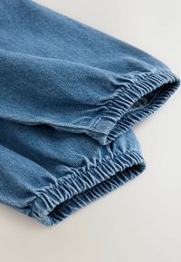 Poignets élastiqués en denim bleu sur un pantalon, montrant la texture et les détails de couture sur un fond clair.
