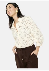 Blusa crema button-up con intricati motivi ritagliati e colletto, abbinata a pantaloni marroni scuri con cordoncino. Tessuto morbido e leggero.