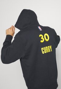 Personne portant un hoodie noir avec le logo de la NBA, le numéro "30" en jaune et le texte "CURRY" dans le dos, tirant la capuche sur sa tête.