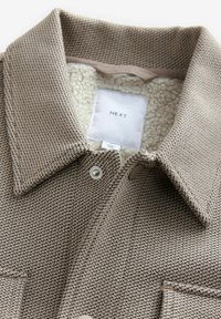 Veste beige en tissu texturé avec boutons-pression, col large et doublure en polaire blanche portant l’étiquette "NEXT" sur la couture intérieure du col.