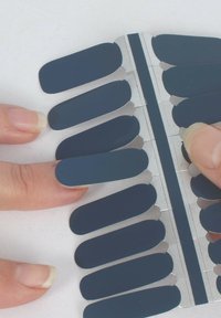 Nail wraps in a dark navy color arranged on a metal strip, with displayed fingers applying one wrap. Smooth texture, oval shape.
