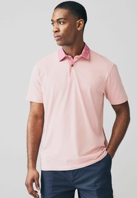 Ljusrosa pikétröja med krage i mörkrosa mönster och en svart knapp vid halsringningen. Kort ärm och slät textur.