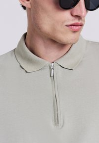 Polo shirt zippé vert clair avec une texture douce et un col ; il présente une courte fermeture éclair sur le devant et un design ajusté, porté avec des lunettes de soleil noires.