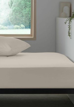 Lit simple avec une literie beige et un oreiller à côté d'une fenêtre montrant des arbres verts, avec une étagère blanche et une plante suspendue à proximité.