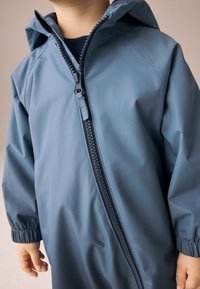 Mėlyna neperšlampama striukė su priekiu esančia užtrauktuku, elastinėmis riešo apyrankėmis ir kapišonu. Pastebimos lygios tekstūros ir lengvo dizaino savybės.