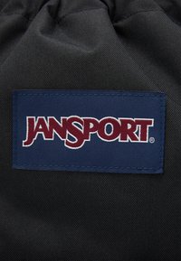 Sac en tissu noir avec une pièce en bleu marine affichant le logo "JANSPORT" en broderie blanche et bordeaux. Surface texturée, design simple.