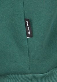 Sudadera verde con un tejido texturizado. Una etiqueta negra con la palabra "PROHIBIDO" está cosida en la costura lateral. Puños y dobladillo acanalados.