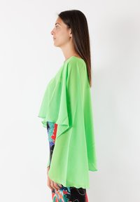 Blusa di un verde brillante, fluida, con maniche larghe e sovrapposte. Tessuto trasparente, design semplice e tessuto leggero. Abbinata a pantaloni colorati e con motivi.