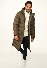 Langer olivgrüner Steppmantel mit Kapuze, Reißverschluss und Seitentaschen, getragen über einem cremefarbenen Zopfmuster-Pullover und marineblauen Hosen. Weiße Sneaker.