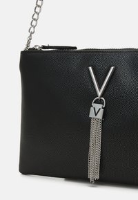 Melnas teksturētas clutch somiņas ar ķēdes siksnu, ar sudraba "V" aksesuāru un uzkausēšanas detaļu ar vairākām ķēdēm. Augšā ar rāvējslēdzēja aizdari.