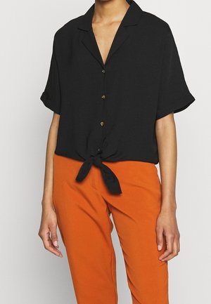 Czarna koszula z krótkim rękawem i kołnierzykiem z wycięciem, z wiązanym przodem i złotymi guzikami. W zestawie z pomarańczowymi spodniami dla kontrastu.