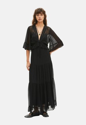 Donna che indossa un lungo abito nero trasparente con maniche svasate, una scollatura profonda a V, una collana nera di perline e scarpe nere slip-on con occhielli argentati.