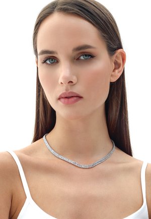Giovane donna con capelli castani lisci indossa una collana di diamanti taglio baguette color argento e una canottiera bianca a spalline sottili su sfondo bianco.