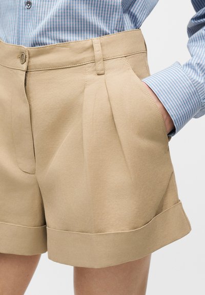 Μπεζ σορτς με ψηλή μέση, γυρισμένα μανσέτα, πλαϊνές τσέπες, πιέτες και κλείσιμο με κουμπί, φορεμένα με μπλε ριγέ πουκάμισο μακρυμάνικο.