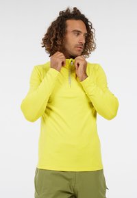 Man met krullend haar in een felgele top met lange mouwen, die de halve rits van zijn kraag aanpast, en olijfgroene broek voor een witte achtergrond.
