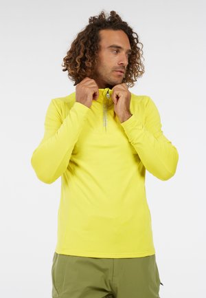 Man met krullend haar in een felgele top met lange mouwen, die de halve rits van zijn kraag aanpast, en olijfgroene broek voor een witte achtergrond.