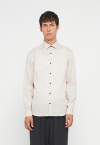 Young man wearing a light beige button-up long-sleeve shirt and dark trousers, standing against a plain white background.
