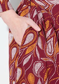 Burgundské šaty s paisley vzorem v bílé a oranžové barvě, s boční kapsou a záhyby pro zvýraznění textury.