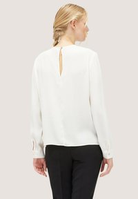 Weißes Langarm-Bluse mit einem Schlitzdetail am Rücken und Knopfverschluss. Glattes Material, lockere Passform und leicht verjüngte Manschetten. Schwarze Hose sichtbar.