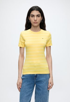 Femme aux longs cheveux foncés portant un t-shirt jaune à manches courtes rayé et un jean bleu, debout devant un fond uni.