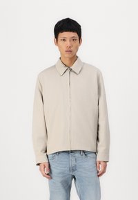 Beige lightweight jacket with a classic collar, front zipper, and long sleeves, paired with blue denim jeans, against a plain background.