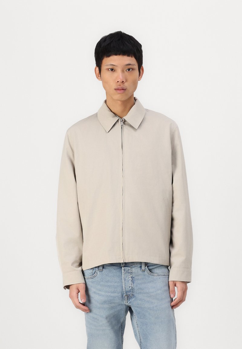 Beige lightweight jacket with a classic collar, front zipper, and long sleeves, paired with blue denim jeans, against a plain background.