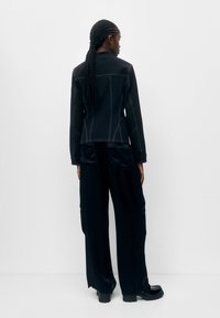 Veste noire avec couture blanche, coupe ajustée, col montant. Associée à un pantalon noir ample et brillant, et des chaussures noires épaisses.