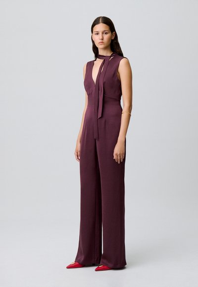 Mono sin mangas burdeos con escote en V profundo y detalle de lazo, confeccionado en tejido suave y con pantalones de pierna ancha. Combinado con zapatos de tacón en punta rojos.