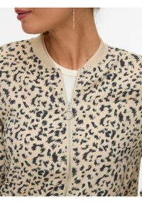 Femme portant une veste zippée beige et noire à motifs tachetés avec un extracteur de fermeture éclair en forme d'anneau et une fine boucle d'oreille pendante.