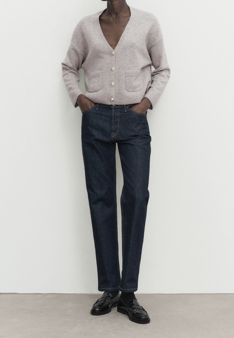 Personne portant un cardigan boutonné gris clair avec des poches, un jean droit bleu foncé et des mocassins noirs, debout les mains dans les poches.
