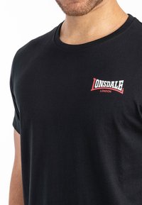 Musta puuvillane T-särk, millel on ümber kaela lõige ja lühikesed varrukad. Vasakul rinnataskul on silmapaistev punane ja valge "Lonsdale London" logo.