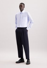 Lichtblauw overhemden met een borstzak, gecombineerd met losvallende marineblauwe broek en zwarte veterschoenen. Gladde stofstructuur.