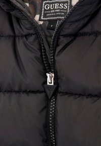 Schwarze Daunenjacke mit einem Reißverschluss auf der Vorderseite. Sie besitzt eine gesteppten Struktur und ein dreieckiges Logoschild in der Nähe des Reißverschlusses. Innenfutter im Camouflage-Muster.