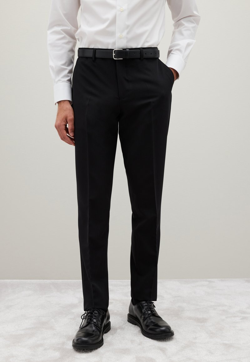 Homme portant un pantalon noir sur mesure, une ceinture noire, des chaussures en cuir noires, et une chemise blanche à manches longues boutonnée, debout sur un tapis clair.