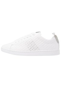 Zapatilla blanca con parte superior texturizada que presenta un logotipo repetido. Punta redonda, acabado suave, suela plana y un acento gris en el talón.