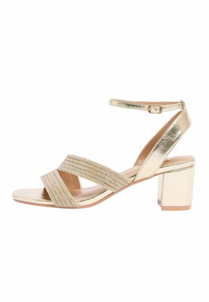 Zlatá sandála s blokovým podpätkom, remienkom okolo členku a krížovo preloženými prednými remienkami ozdobenými korálkami na bielom pozadí.