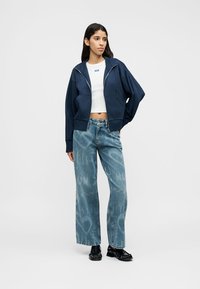 Μπλε ναυτικό κοντό μπουφάν με φερμουάρ, φορεμένο πάνω από ένα λευκό κοντό τοπ, συνδυασμένο με ψηλόμεσα ανοιχτό μπλε τζιν με διακριτή υφή. Μαύρα παπούτσια ολοκληρώνουν την εμφάνιση.