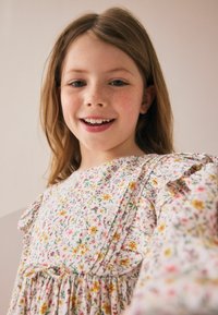 Blumenkleid mit weißem Hintergrund, das farbenfrohe Blumen in Pink, Gelb und Grün zeigt. Mit Rüschensäumen und cinchtem Taillenbereich.