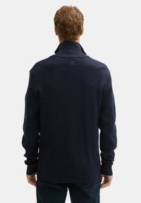 Granatowy sweter wykonany na drutach z wysokim kołnierzem, ściągaczami przy rękawach oraz kwadratową metką z tyłu. Teksturowana tkanina i luźny krój.