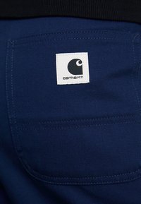 Gros plan sur une poche en tissu bleu avec un patch logo Carhartt carré blanc cousu au centre de la poche.