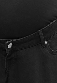 Nærbillede af linning på sorte jeans, der hviler på en sort stofbaggrund, som viser knap, bæltestrop og detaljer på forlomme.