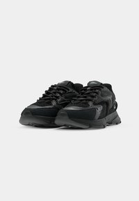 Paire de chaussures de sport noires avec mesh, accents en cuir et semelles épaisses, présentées sur un fond gris clair uni.
