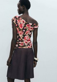 Top floreale marrone con motivi rosa e verdi, design off-shoulder. Abbinato a pantaloni corti grigio scuro fino alla caviglia e accessori chunky.