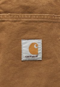 Καφέ ύφασμα με ορατές ραφές και μια λευκή τετράγωνη ετικέτα με πορτοκαλί λογότυπο Carhartt και κείμενο ραμμένα στην επιφάνεια.