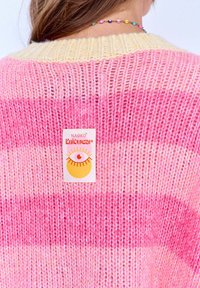 Przesadny sweter dzianinowy w różowo-kremowe paski. Posiada ściągacz przy dekolcie oraz metkę z motywem oka. Miękka faktura i swobodny styl.