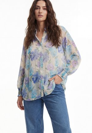 Woman with long wavy hair wearing a sheer, long-sleeve floral blouse in blue and purple tones paired with blue jeans.