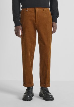 Pantaloni in corduroy marroni con righe verticali, dal taglio sartoriale e con orli arrotolati, abbinati a stivali neri con lacci.