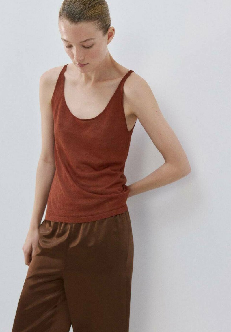 Femme aux cheveux attachés portant un haut sans manches couleur rouille et un pantalon en satin marron, regardant vers le bas avec une main derrière le dos.