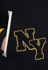 Tengerészkék pulóver texturált sárga-fekete "NY" felvarróval a mellkason, bordázott szegélyekkel és polárszerű anyagból.