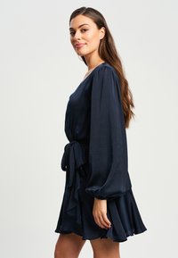 Mornarsko modra obleka z dolgimi balonskimi rokavi, prilegajočim pasom in nabranim robom. Narejena iz lahkega, teksturiranega materiala.